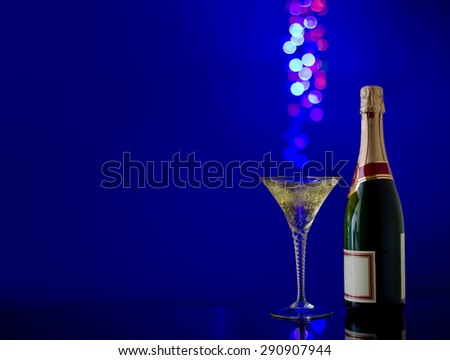 Champagne glass with sparkling bokeh light effect in background and a full bottle of unopened champagne or sparkling wine with clean label. Dark blue background with vignetting in corners.
