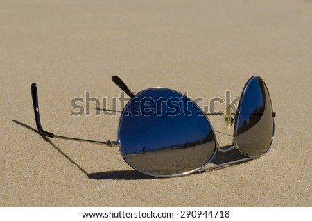 Details of a pair of dark tinted sunglasses on beach sand. Lenses have dark mirror-like reflective surface and reflection of beach and blue sky can be seen.