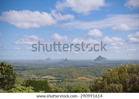 Panoramic view of the Glasshouse Mountains, Sunshine Coast, Queensland, Australia. Top two third of image is blue sky with white clouds. Green vegetation and forest area in foreground.