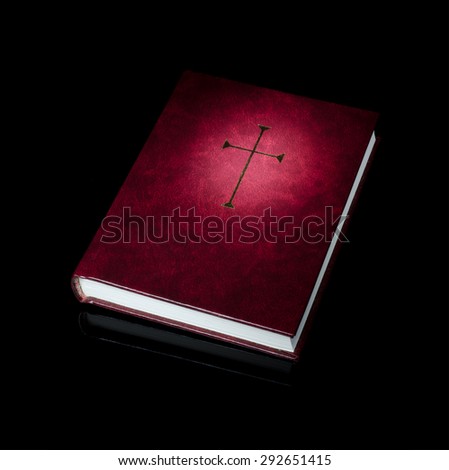 Low key shot burgundy prayer book with gold cross on it. Golden cross has light projected on it. Book has slight reflection on black background.