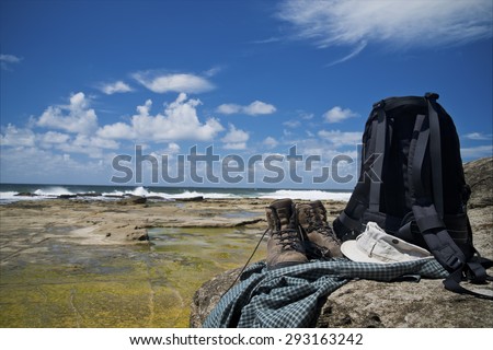 A pair of old hiker\'s boot, hat, sunglasses, shirt and backpack on a rock by the ocean. Waves crashing on rocky seashore. Blue sky with some clouds.