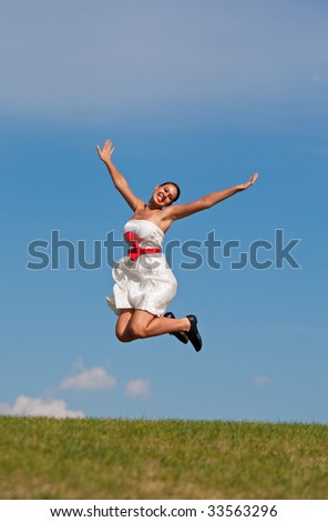 A beautiful girl in a white dress jumping for joy on a grassy, green hill, on a warm summer day.