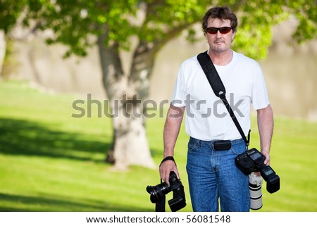 A professional photographer armed with two cameras, walking on the grassy shores of a river.