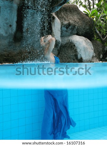 Young blond woman under a waterfall in the pool with an evening gown on - trash the dress trend.