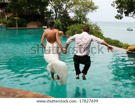 Happy bride and groom jump into the swimming pool during a trash the dress photo shoot.