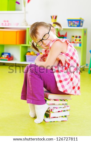 Cute little girl in eyeglasses sitting on stack of books at home