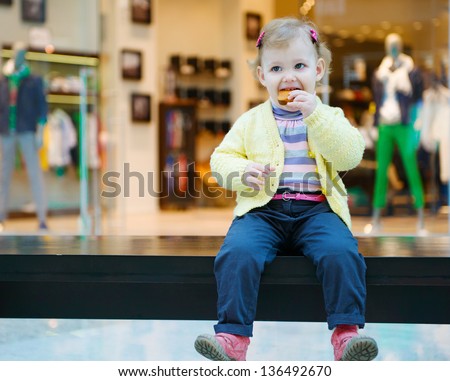 Cute little girl eating biscuit cake on bench in mall