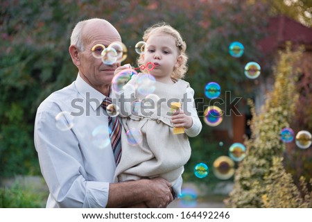 Happy grandfather and cute curly granddaughter blowing soap bubbles