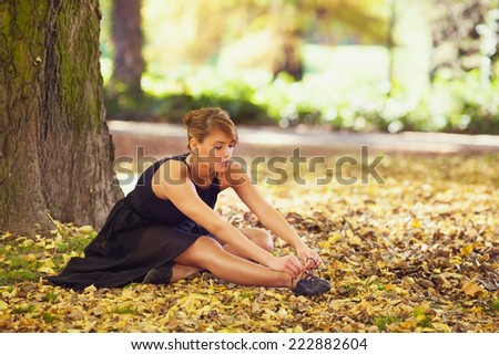 Beautiful ballerina tying shoelace sitting among autumn leaves