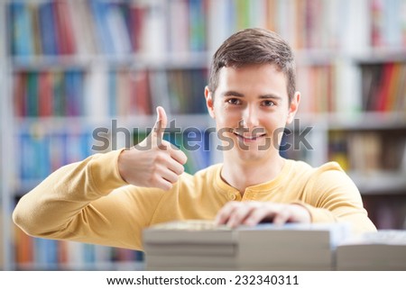 Young man standing among books in the library showing ok sign