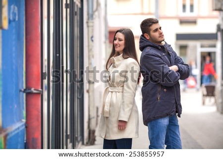 Young woman looking at store window and bored young man standing behind her back with his arms crossed waiting for her.