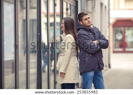 Young couple standing in front of the boutique. Girl looking at store window and bored young man standing behind her back with his arms crossed waiting for her.