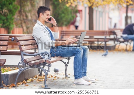 Young man talking on his mobile phone while sitting on the park bench with laptop on his lap