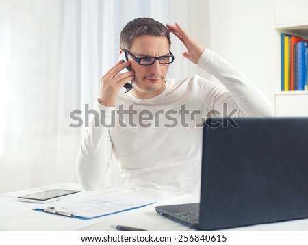 Portrait of young businessman scratching his head while working at home using computer and mobile phone.