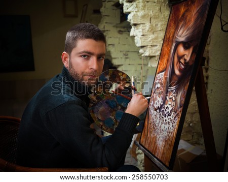 Artist sitting in his studio holding cigarette and paintbrushes working on oil painting portrait of beautiful young woman.