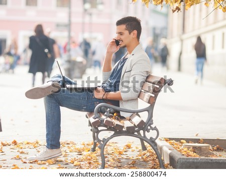 Handsome young man sitting on the bench in the city using laptop and talking on the phone.