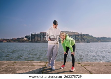 Young woman having a break from running standing with her hands on knees. Young Man next to her using mobile phone.