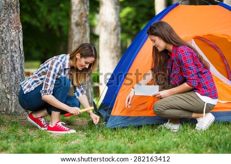 Young woman putting up a tent with her friend helping her reading a manual