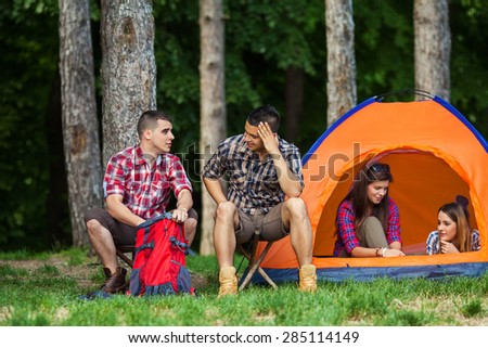 Two young men talking and searching for something in backpack while sitting in front of the tent. Two young women relaxing and talking in a tent
