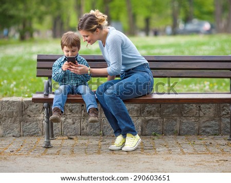 Cute little boy sitting on a park bench with his mother and playing with mobile phone