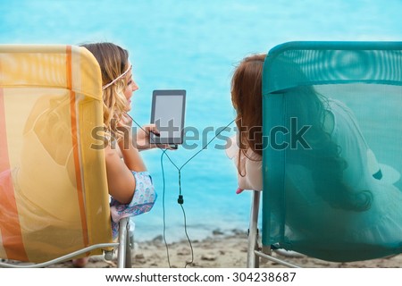 Rear view of two girls listening to music on digital tablet while sunbathing on the beach