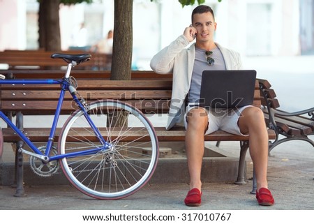 A handsome businessman working on his laptop and talking on the phone while sitting on a park bench