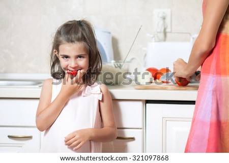 Cute little girl eating tomato while helping her mother in the kitchen