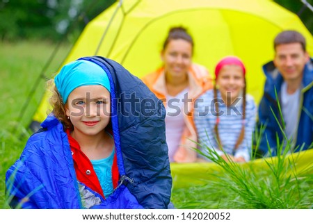 Young girl in a sleeping bag with her family near tent in camping on the nature
