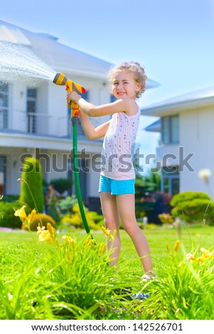 Little gardener girl watering flowers on the lawn near cottage. Vertical view