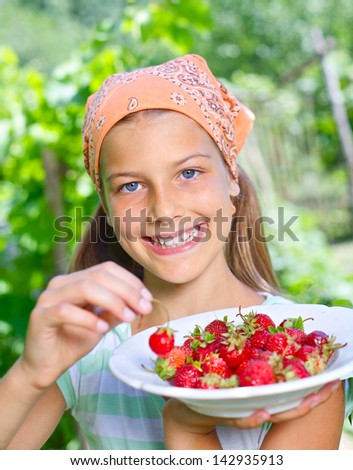 Beautiful little smiling girl eating strawberries in garden. Vertical view