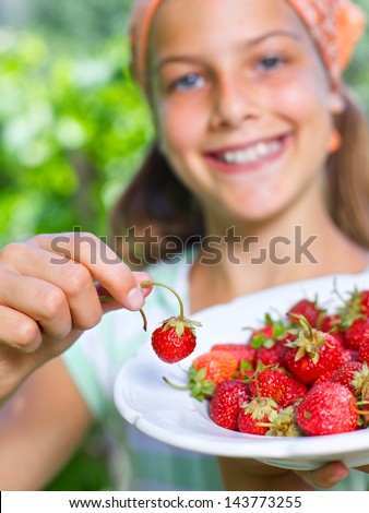 Beautiful little smiling girl eating strawberries in garden. Focus of strawberries. Vertical view