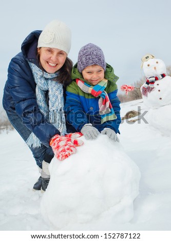 Happy beautiful boy with mother building snowman outside in winter time