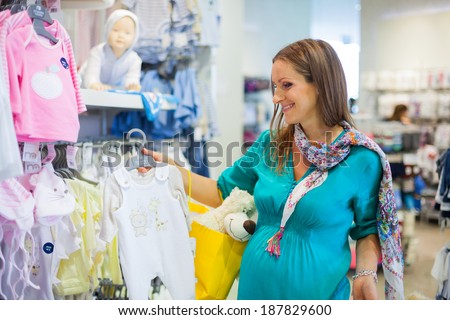Young pregnant woman choosing newborn clothes at baby shop store