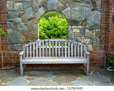 A wooden bench on a patio with stonewall blocking the view into the garden.  A small arch window in the stonewall allows a small view into the garden beyond.