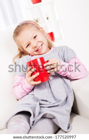 Cute little girl drinking tea from a red cup at home.