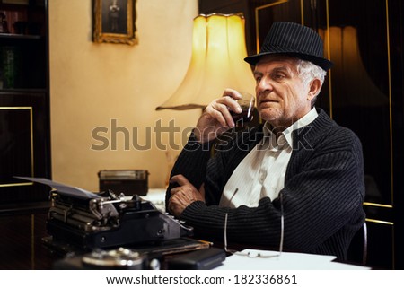 Retro Senior man writer with hat, sitting at the desk with glass of whiskey and thinking.