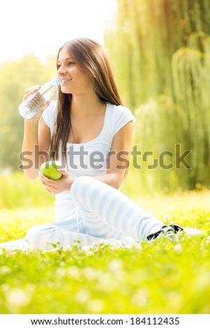 Smiling young woman holding an apple, drinks water from a bottle and enjoying in the nature.
