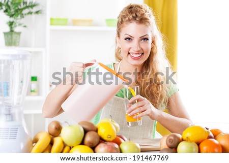 Beautiful young woman pouring orange juice at glass in a kitchen. Looking at camera.