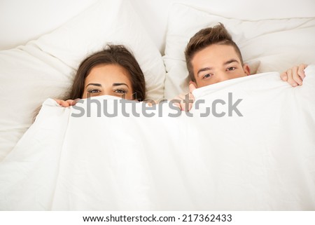 Funny couple in bed looking and peeking over sheets smiling.