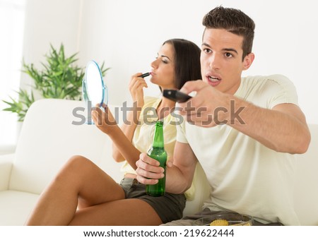 Young man sitting on sofa and watching football on tv. His girlfriend bored and make up lipstick on mouth.
