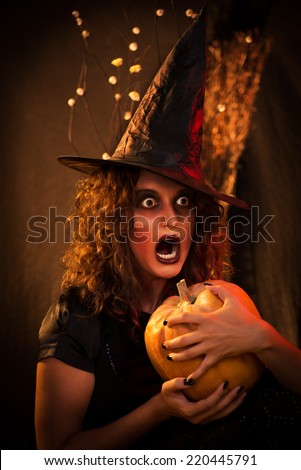 Young woman with scared face dressed like a witch. She wears dark clothing and holding a pumpkin in hands.