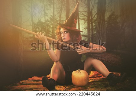 Young woman dressed like a witch. She is sitting in dark clothing with broom and pumpkin.