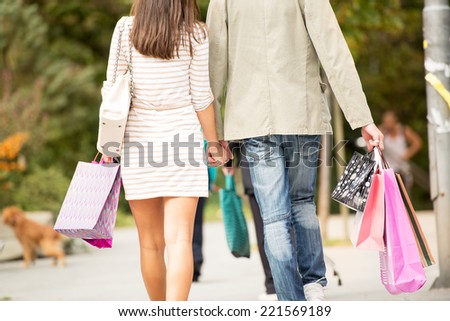 Couple in love walking through town after shopping, holding hands and carrying a bunch of shopping bags, backs to the camera.