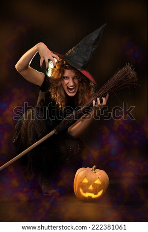 Young woman dressed like a witch. She is in dark with broom and pumpkin.