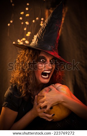 Young woman with evil face dressed like a witch. She wears dark clothing and holding a pumpkin in hands.