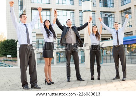 Small group of business people outside their company, holding hands raised celebrate business success.