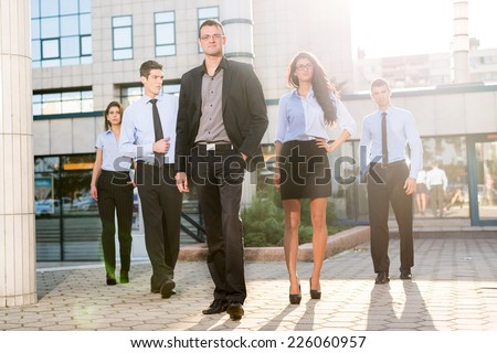 Young businessman, elegantly dressed with his hand in his pocket, standing proudly with his team of young businesswomen and businessmen in front of office building illuminated backlit sun.