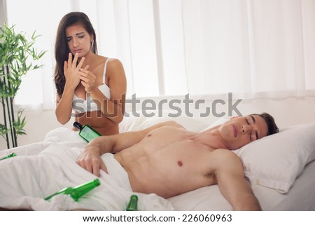 A young woman had just woke up next to a naked man who sleeps next to pile of empty bottles with disappointment and sadness looking at the wedding ring on her hand.