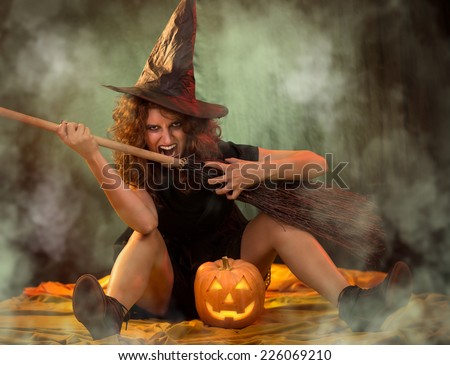 Young woman dressed like a witch. She is sitting in dark clothing with broom and pumpkin.