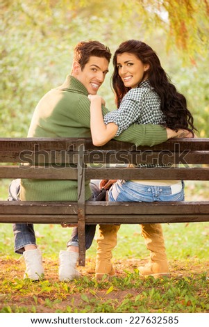 Young heterosexual couple hugging sitting on a park bench, behind their back looking at the camera with a smile on their faces.